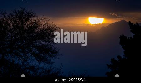A dramatic sunset behind mountains, partially obscured by clouds. Silhouetted trees frame the scene, creating a serene and moody atmosphere Stock Photo