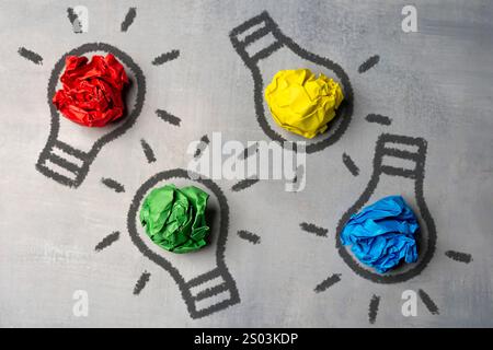 A flat lay image of multiple lightbulb drawings on a chalkboard with crumpled pieces of paper in red, yellow, green, and blue. Brainstorming concept. Stock Photo
