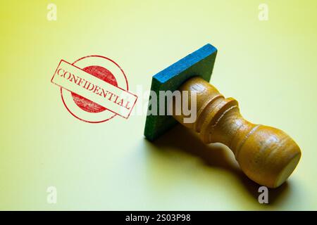 A close-up image of a red "CONFIDENTIAL" stamp imprint on a yellow background, with a wooden stamp. Stock Photo