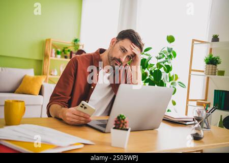 Man holding phone and sitting at desk with laptop in bright home interior Stock Photo