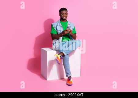 Relaxed young man with a bright smile sits against a vibrant pink background, casually using his phone Stock Photo