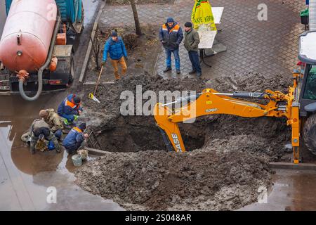 Ukraine, Romny, December 24, 2024: A group of workers repair a damaged pipeline using heavy equipment, including a backhoe loader and a sewage truck. Stock Photo