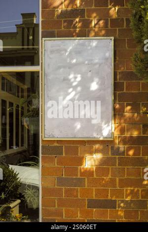 A blank white signboard mounted on a brick wall, with natural light creating dappled shadows, next to a window reflecting part of a building and green Stock Photo