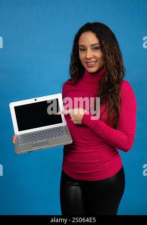Smiling young woman pointing at a laptop with blank screen on a blue background Stock Photo