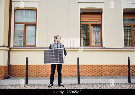 Man holding solar panel, stands in front of old building. Guy in a black hoodie and navy blue quilted vest. Aged architecture contrasts with modernity of renewable energy. Stock Photo