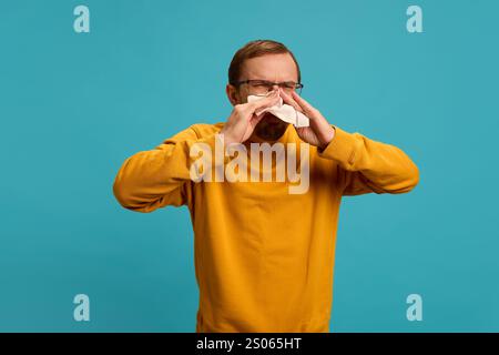 Young man dressed casually in glasses suffering from sneezing, blows his nose into napkin against blue studio background. Stock Photo