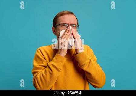 Young man dressed casually in glasses suffering from sneezing, blows his nose into napkin against blue studio background. Stock Photo