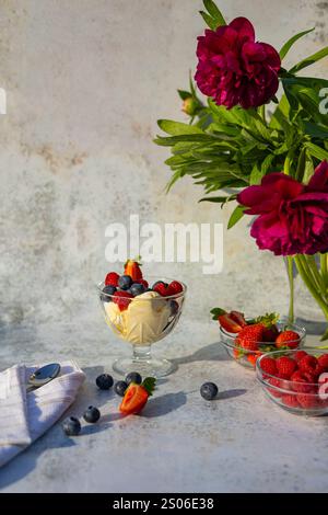 Vanilla ice cream served in a glass bowl, garnished with fresh berries, placed on a rustic table with vibrant red peonies in soft natural sunlight Stock Photo