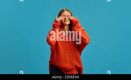 Young woman in orange sweater closing her eyes appearing in discomfort isolated on a blue background. Allergy. Stock Photo