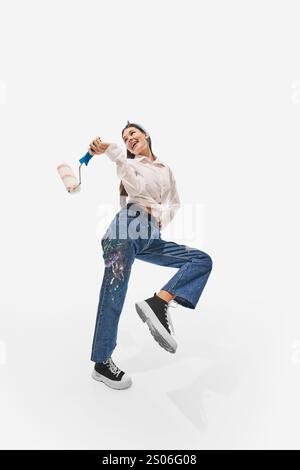 Painter wearing white shirt and paint-splattered jeans, holding paint roller, smiling vibrantly while posing against white studio background. Stock Photo