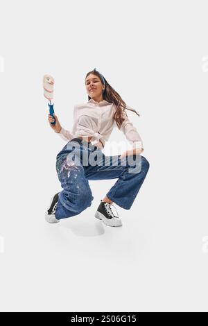 Young woman in white shirt and colorful jeans leans forward holding paint roller, expressing creativity against white studio background. Stock Photo