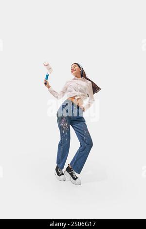 Woman dressed in white shirt and paint-splattered jeans holds paint roller and dancing with joy against white studio background. Stock Photo