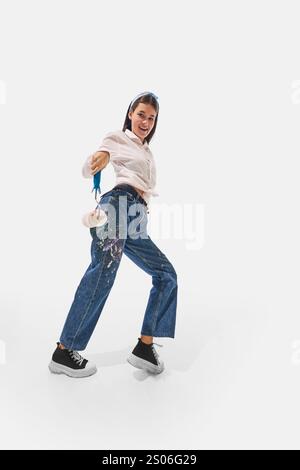 Painter in white shirt and colorful jeans leaning backward, raising paint roller playfully with joyful expression against white studio background. Stock Photo