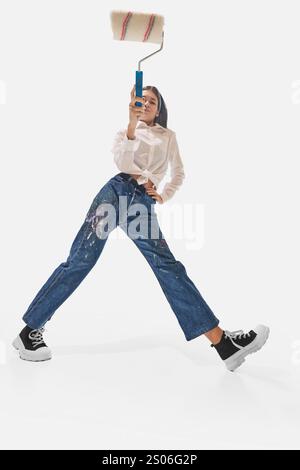 Energetic woman with paint roller jumps enthusiastically in denim pants splattered with paint, highlight style against white studio background. Stock Photo