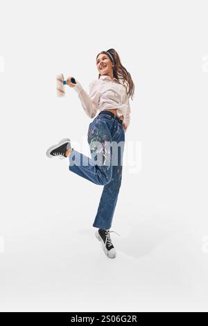 Energetic woman with paint roller jumps enthusiastically in denim pants splattered with paint, highlight style against white studio background. Stock Photo