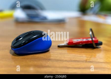 A blue and black computer mouse sits next to a set of keys on a wooden table. The background features scattered office supplies, indicating a workspac Stock Photo