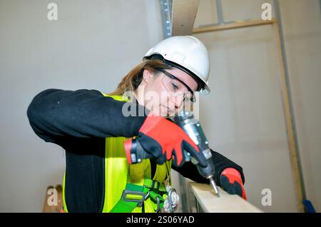 Female construction worker using power drill, mounting plasterboard on a construction site, wearing safety helmet, glasses and gloves Stock Photo