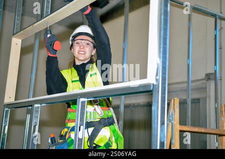 Female construction worker assembling plasterboard on metal frame using electric screwdriver, wearing safety helmet and harness Stock Photo