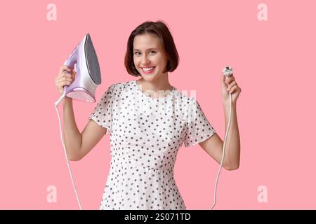 Smiling young woman holding iron on pink background Stock Photo