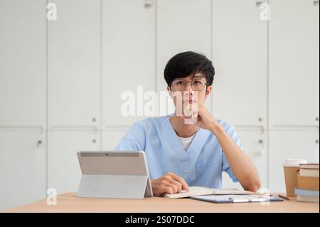 A smart Asian male medical student in scrubs, wearing eyeglasses, is focused on listening to a professor teaching in the classroom while sitting at a Stock Photo