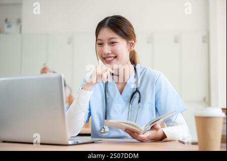 A pretty and smart Asian female medical student in scrubs is focused on reading medical cases on her laptop while attending a lecture with a professor Stock Photo