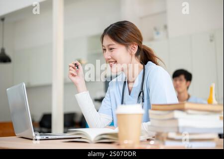 A pretty and smart Asian female medical student in scrubs is focused on reading medical cases on her laptop while attending a lecture with a professor Stock Photo