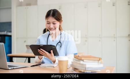 A pretty and smart Asian female medical student in scrubs is focused on reading medical cases on her tablet while attending a lecture with a professor Stock Photo