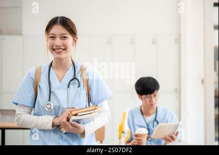A portrait of a beautiful, smart Asian female medical student in scrubs standing in her classroom, carrying medical textbooks, and smiling at the came Stock Photo