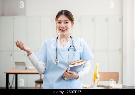 A portrait of a beautiful, smart Asian female medical student in scrubs standing in her classroom, carrying medical textbooks, and smiling at the came Stock Photo