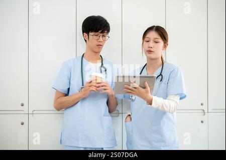 Two smart Asian male and female doctors or medical students in scrubs are looking at a tablet and discussing the medical cases on the screen in the lo Stock Photo