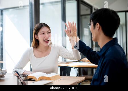 Two cheerful young Asian college students are giving each other a high five to celebrate their exam success while studying together in a library or co Stock Photo