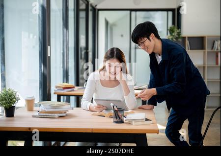 Two positive young Asian college students are looking at a tablet screen and discussing a topic for the exam while studying and preparing together at Stock Photo