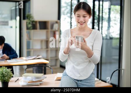 A portrait of a beautiful, smiling young Asian female college student in casual clothes reading texts on her smartphone while standing in a library. p Stock Photo