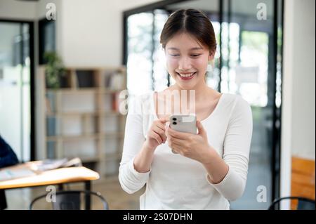 A portrait of a beautiful, smiling young Asian female college student in casual clothes reading texts on her smartphone while standing in a library. p Stock Photo