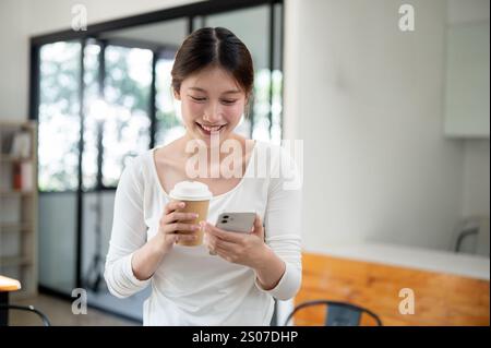 A portrait of a beautiful, smiling young Asian woman in casual clothes holding a hot drink cup and reading texts on her smartphone while standing in a Stock Photo