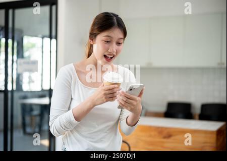 A portrait of an excited young Asian woman in casual clothes holding a cup of hot coffee and reading texts on her smartphone with a surprised expressi Stock Photo