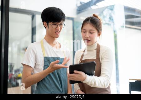 Two Asian coffee shop staff members are discussing the menu on a tablet while working in their coffee shop during the daytime. small business, part-ti Stock Photo