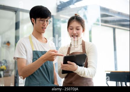 Two Asian coffee shop staff members are discussing the menu on a tablet while working in their coffee shop during the daytime. small business, part-ti Stock Photo