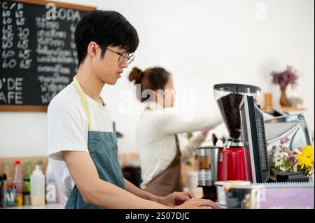 A friendly young Asian male coffee shop staff member or barista is working on a computer at the cashier checkpoint at the counter, working in a coffee Stock Photo