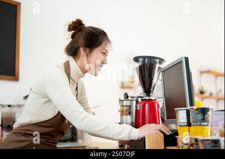 A friendly, smiling young Asian female coffee shop staff member or barista is working on a computer at the cashier checkpoint at the counter, working Stock Photo