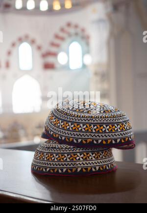 Three embroidered men's Turkmen taquiyahs or tahya with geometric white and orange designs on black fabric. They're stacked on a wooden railing in the Stock Photo
