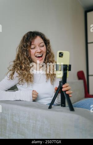 Young woman with curly hair laughing while recording a video blog using her smartphone on a tripod, sitting on a sofa in her living room, creating engaging content for her online audience Stock Photo