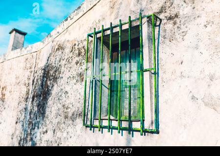 Rectangular window with greenish glass panes is set in weathered, white wall. Window is protected by metal grille with vertical and horizontal bars, w Stock Photo