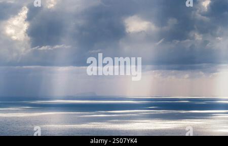 A serene seascape featuring calm waters reflecting sunlight, with dramatic clouds overhead and rays of light breaking through, creating a tranquil atm Stock Photo
