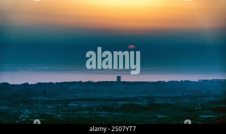 A serene sunset over a coastal landscape, with the sun partially submerged in the horizon. The sky transitions from orange to deep blue, reflecting on Stock Photo