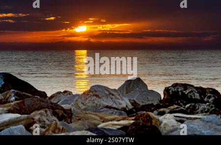 A serene sunset over the ocean, with vibrant orange and yellow hues reflecting on the water's surface. In the foreground, smooth rocks are scattered a Stock Photo