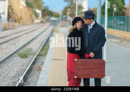 A couple in love at the train station, waiting for the train for their honeymoon, carrying suitcases and vintage clothes. Stock Photo