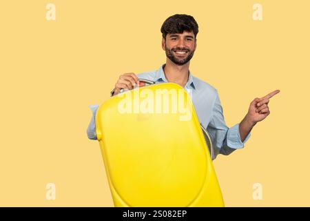Young businessman holding yellow suitcase and pointing with index finger to the side on yellow background Stock Photo
