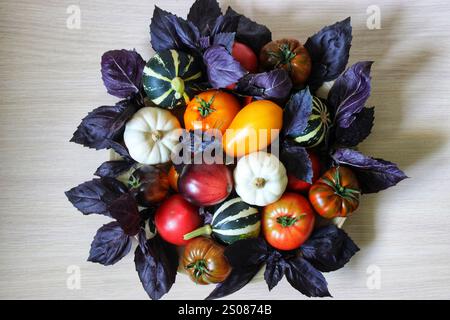 Freshly harvested heirloom tomatoes, pumpkins, and purple basil leaves arranged on a wooden surface, creating a vibrant and colorful display Stock Photo