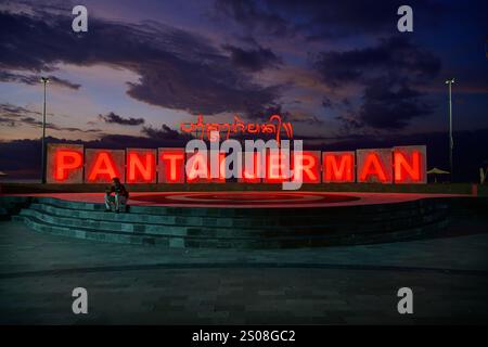 Jerman Beach, Bali, Indonesia - November 7 2024: Man Sitting, Reading on his Smartphone by the Illuminated Sign of Pantai Jerman Beach. Stock Photo